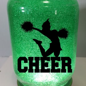 Pu&ograve; includere: Un barattolo di vetro verde con glitter, una silhouette nera di una cheerleader con pompon e la parola "CHEER" in lettere nere.