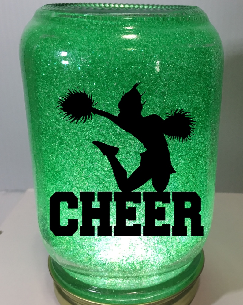 Pu&ograve; includere: Un barattolo di vetro verde con glitter, una silhouette nera di una cheerleader con pompon e la parola "CHEER" in lettere nere.