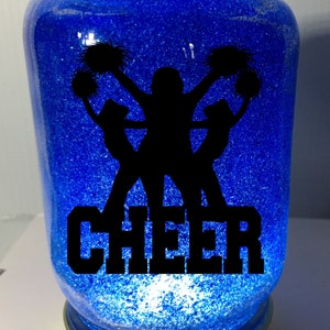 Pu&ograve; includere: Un barattolo di vetro blu con glitter e una silhouette nera di cheerleader con pompon. La parola "CHEER" &egrave; scritta in lettere nere sotto le cheerleader.