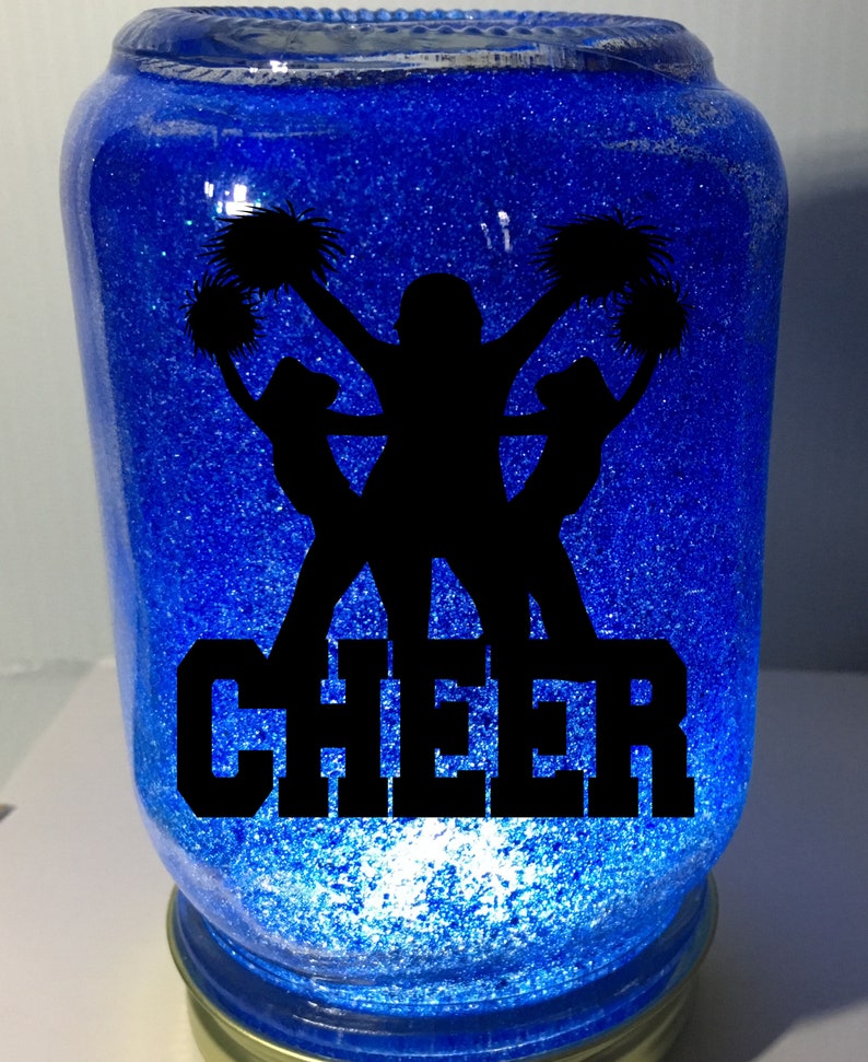 Pu&ograve; includere: Un barattolo di vetro blu con glitter e una silhouette nera di cheerleader con pompon. La parola "CHEER" &egrave; scritta in lettere nere sotto le cheerleader.