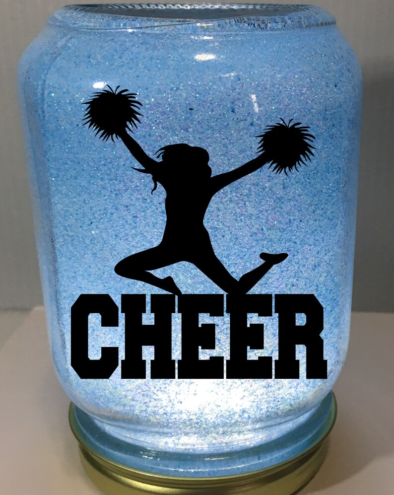 Pu&ograve; includere: Un barattolo blu con glitter con la silhouette di una cheerleader che tiene pompon e la parola "CHEER" in lettere nere.