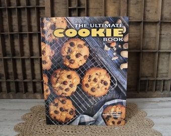 May include: The Ultimate Cookie Book cookbook cover features a close-up of a variety of cookies on a wire rack. The cookies are chocolate chip, peanut butter, and other varieties. The book title is in large, yellow letters on a blue background.