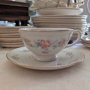 May include: A white teacup and saucer set with a floral design in pink and yellow. The teacup and saucer have gold trim. A stack of plates and other teacups are in the background.