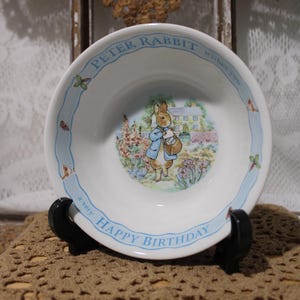 May include: White ceramic bowl featuring a Peter Rabbit illustration. The bowl has a light blue border with the text "PETER RABBIT wishes you a very HAPPY BIRTHDAY". The center shows Peter Rabbit in a blue coat.