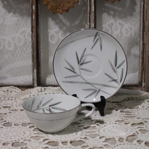 May include: A white teacup and saucer set with a bamboo leaf design. The cup has a handle and a silver rim, and the saucer has a matching design. The set is displayed on a white lace tablecloth.