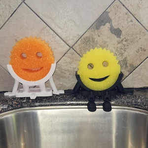 May include: Two smiling sponge holders, one orange and one yellow, sit above a stainless steel sink. The orange sponge is held by a white stand, while the yellow sponge is held by a black stand. Both have a smiley face design.