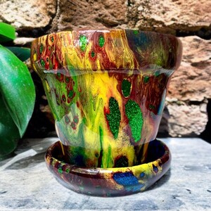 May include: A brightly coloured ceramic plant pot and saucer set. The pot displays an abstract design with streaks of yellow, red, green, and brown, with green glitter accents. The saucer echoes the pot's design, creating a cohesive look.