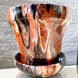 May include: A black, orange, and white ceramic planter with a matching saucer. The planter has a swirled, abstract design with gold glitter accents.