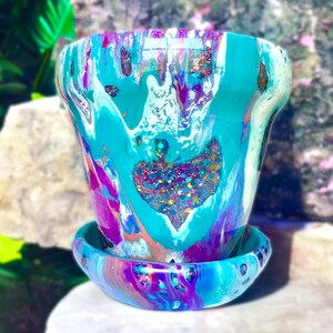 May include: A teal, purple and white ceramic planter with a matching saucer. The planter has a heart-shaped design with glitter embedded in the centre.
