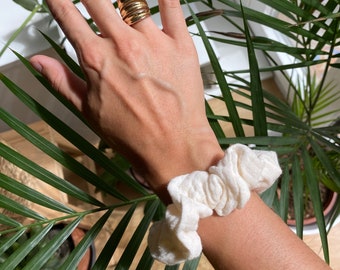 May include: A gold ring with multiple bands stacked on top of each other. The ring is worn on a person's finger. A white scrunchie is worn on the person's wrist.