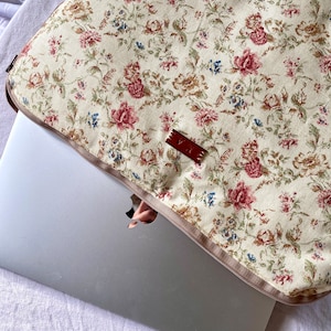 Può includere: Una custodia per laptop beige con un motivo floreale con fiori rossi, rosa e blu. La custodia ha una chiusura a zip e un'etichetta in pelle marrone con la scritta "VUE" su di essa.