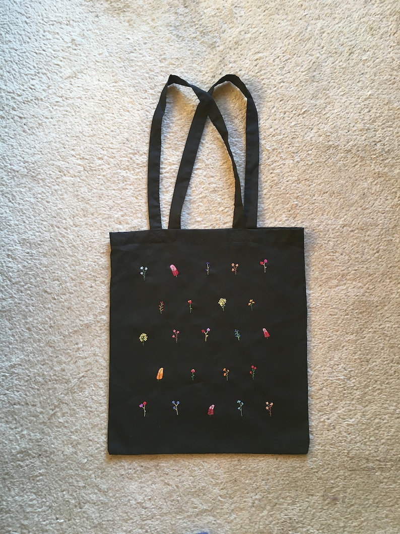 以下が含まれることがあります： 黒いトートバッグに、カラフルな花と葉が刺繍されています。