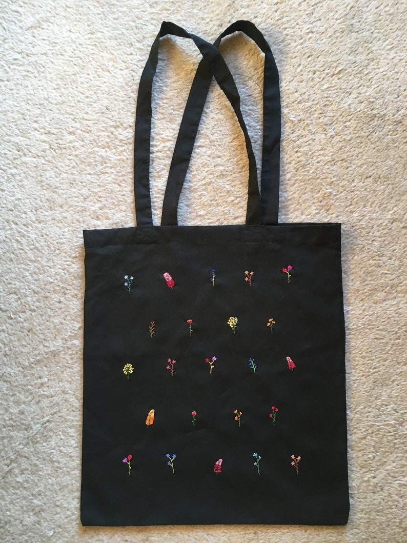 以下が含まれることがあります： カラフルな刺繍の花が施された黒いキャンバス地のトートバッグ。バッグには黒いストラップが2本付いている。