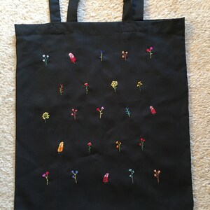 以下が含まれることがあります： 様々な色の刺繍が施された黒いトートバッグ。花はバッグ全体にランダムに散りばめられています。