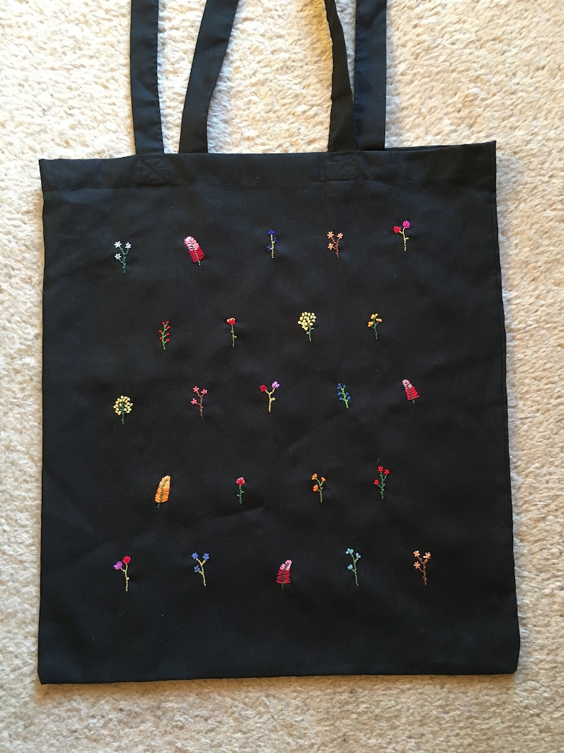 以下が含まれることがあります： 様々な色の刺繍が施された黒いトートバッグ。花はバッグ全体にランダムに散りばめられています。
