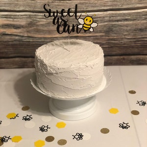 May include: A white cake with white frosting and a black cake topper that says "Sweet as can be" with a cartoon bee. The cake is on a white cake stand and there are white, yellow, and gold confetti on the table.