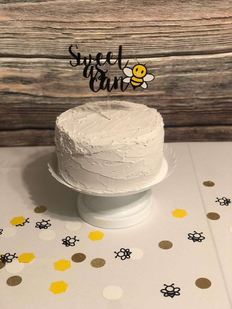 May include: A white cake with white frosting and a black cake topper that says "Sweet as can be" with a cartoon bee. The cake is on a white cake stand and there are white, yellow, and gold confetti on the table.