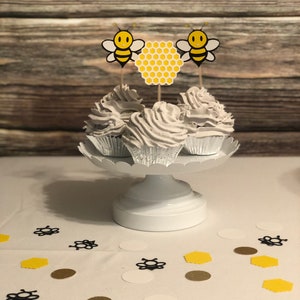 May include: Three white cupcakes with white frosting topped with yellow and black bee and honeycomb cupcake toppers on a white cake stand. The cupcakes are on a white tablecloth with black and gold confetti.