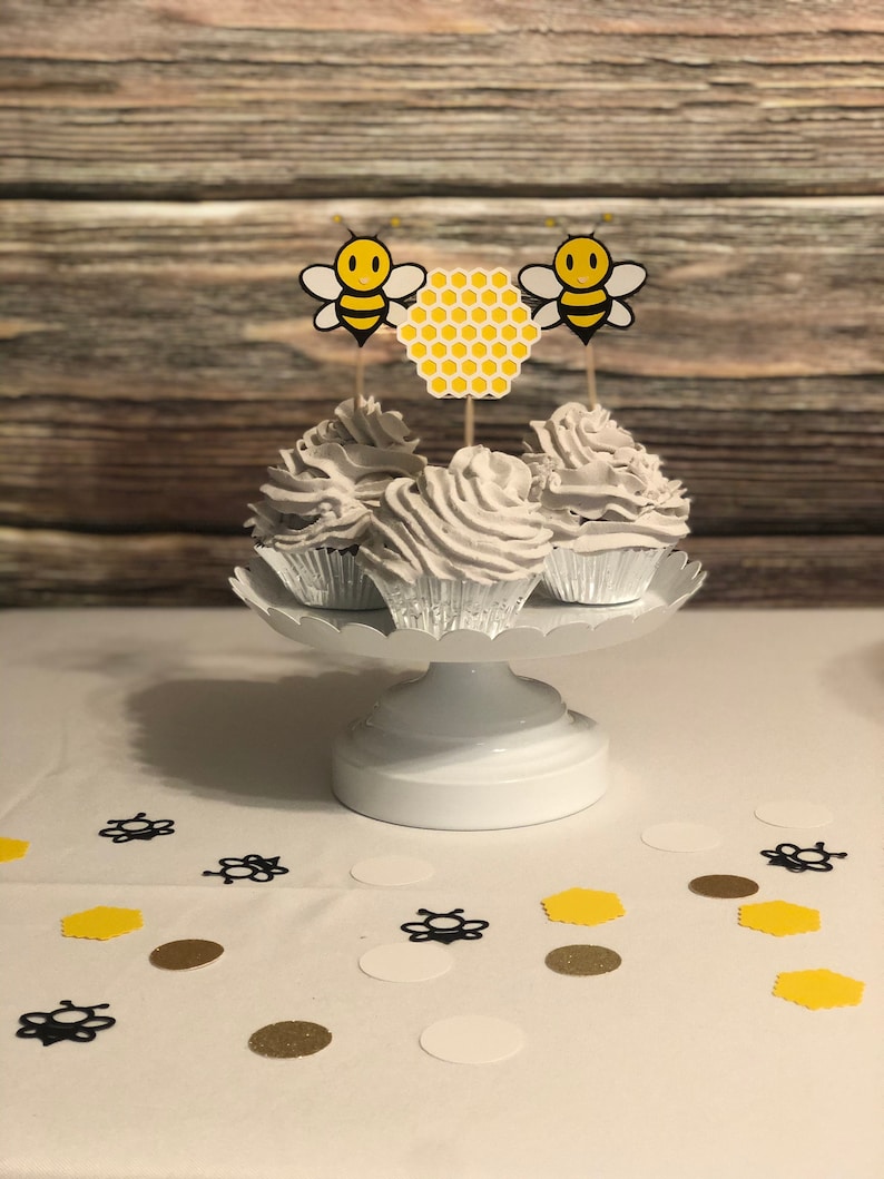 May include: Three white cupcakes with white frosting topped with yellow and black bee and honeycomb cupcake toppers on a white cake stand. The cupcakes are on a white tablecloth with black and gold confetti.