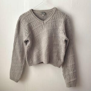 May include: A light beige, open-knit jumper with a V-neck and long sleeves. The jumper is hanging on a wire hanger against a white wall. The knit pattern features diagonal and vertical lines.