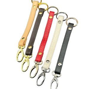 May include: Five leather keychains in various colors: tan, dark brown, red, cream, and black. Each keychain features a metal clasp and a ring for attaching keys. The clasps have gold, silver, and bronze finishes.