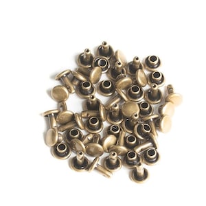 May include: A pile of antique gold metal rivets. The rivets have a round head and a post that is designed to be hammered into a hole.