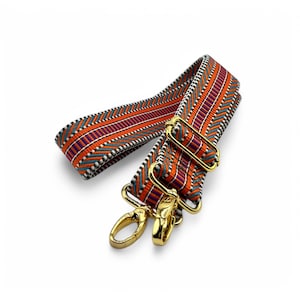 May include: A colorful bag strap with a woven pattern in orange, purple, teal, and white. The strap has gold-colored metal clasps and hardware for attaching to a bag. The strap is designed to be adjustable and is a stylish accessory.