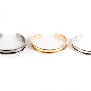 May include: Three metal bracelets in different finishes: black, gold, and silver. Each bracelet has a simple, rounded design with a slightly wider band.