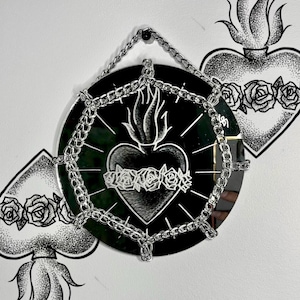 Puede incluir: Espejo redondo negro con borde de cadena plateada y diseño de corazón. El corazón presenta una llama y está rodeado de rosas. Obras de arte adicionales en forma de corazón son visibles en la pared blanca.