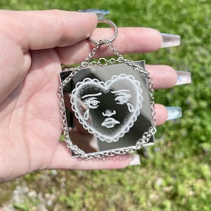 Può includere: Un portachiavi quadrato a specchio con catena e anello portachiavi argentati. Il portachiavi presenta un design a forma di cuore bianco con un volto triste e lacrime. Il disegno è inciso sulla superficie a specchio.