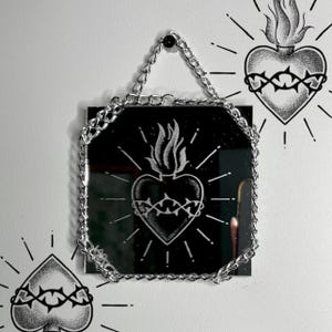 以下が含まれることがあります： 銀色のチェーンで吊るされた、黒い八角形の鏡。鏡には、炎と棘のある白いハートのデザインが施されています。白い壁には、棘のある追加のハートのイラストがあります。