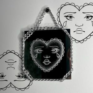 May include: A square mirror with a silver chain hanging from a black screw. The mirror has a white heart-shaped drawing with a face and teardrops. Two other heart drawings are on the white wall.