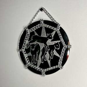 Może przedstawiać: Okrągły, czarny element dekoracyjny ze srebrną obwódką łańcucha i łańcuchem do zawieszania. Element posiada wzór rozbłysku gwiazdy z trzema sylwetkami zwierząt. Element wisi na białej ścianie.