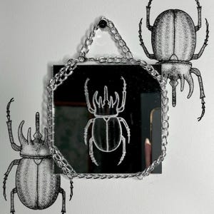 Puede incluir: Espejo cuadrado con borde de cadena plateada, colgado en una pared blanca. El espejo refleja un diseño de escarabajo blanco. Dos ilustraciones de escarabajos adicionales están en la pared, una a cada lado del espejo. Decoración con temática de insectos.
