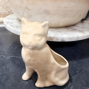 May include: A beige ceramic cat-shaped planter. The planter has an open top for holding plants or other items. The cat is in a seated position with its ears perked up. The planter is on a dark surface.