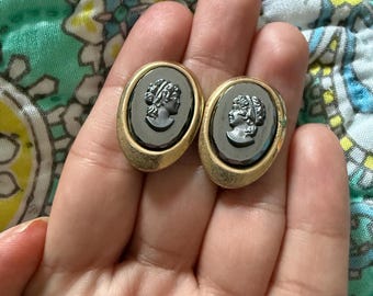 Könnte beinhalten: Ein Paar ovale Cameo-Ohrringe. Jeder Ohrring zeigt ein dunkelgraues Profil in einem goldfarbenen Rahmen. Die Kameen zeigen eine klassische weibliche Figur. Die Ohrringe werden in einer Hand gehalten.