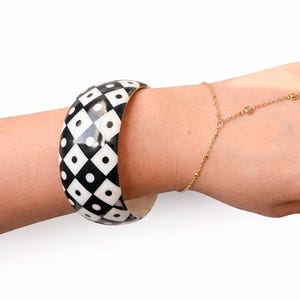 May include: A wide, black and white patterned bangle bracelet. The bracelet features a repeating pattern of black diamonds and white circles. A delicate gold chain bracelet with small clear stones is also visible.