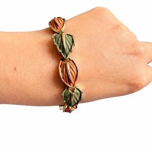 May include: Gold-toned bracelet featuring alternating leaf and bud designs. The leaves are green with gold accents, and the buds are a reddish-orange color. The bracelet is worn on a wrist.