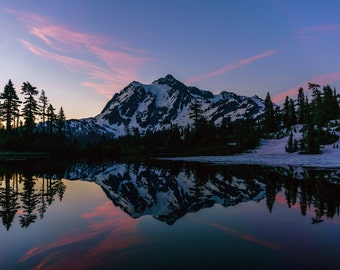 May include: A mountain peak with snow-capped summit reflected in a still lake at sunset. The sky is a vibrant pink and purple with wispy clouds. Silhouettes of trees line the shore.