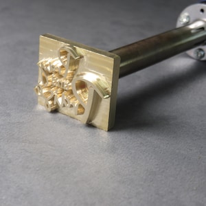 Op de afbeelding: Een gouden metalen stempel met de woorden "Happy Smile Day" erop gegraveerd. De stempel is bevestigd aan een metalen staaf met een blauwe handgreep.