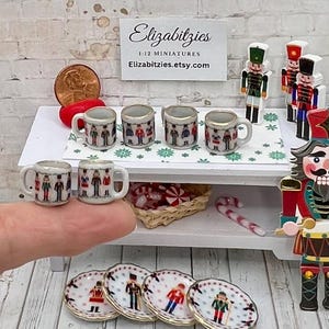May include: Set of six miniature porcelain teacups with a nutcracker design. The cups are white with gold trim and feature a colorful nutcracker pattern. The cups are displayed on a white shelf with a nutcracker figurine and candy canes.