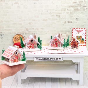 May include: Miniature gingerbread houses with red and white details, snow-covered roofs, and festive decorations. The scene includes small Christmas trees and a "Gingerbread Bakery" sign, all arranged on a white table with a red polka dot pattern.