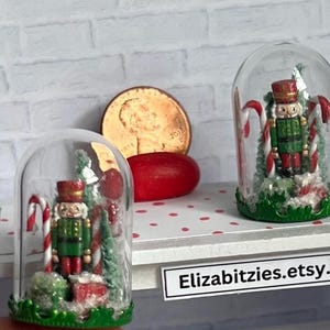 May include: Two miniature glass dome ornaments with a green base. Each dome contains a miniature nutcracker figurine, a small Christmas tree, and candy canes. The domes are sitting on a shelf with a white background and red polka dots.