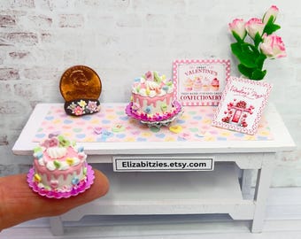 May include: Two miniature pink and white cakes decorated with candy hearts and flowers on a white table with a pink and white Valentine's Day themed tablecloth. The table also has a sign that says "Sweet Valentines Confectionery" and a card that says "Happy Valentine's Day".