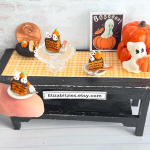 May include: Miniature Halloween-themed display featuring a black table with a yellow checkered tablecloth. Includes a pumpkin, ghost, cake slices, and a framed picture. The cake slices have pumpkin and ghost decorations.