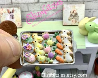 May include: A miniature baking sheet filled with colorful Easter-themed cookies. The cookies are decorated with chicks, carrots, and flowers. The baking sheet is on a white surface.