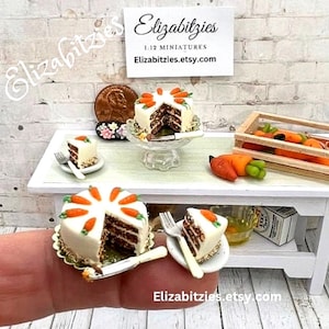 May include: Three miniature carrot cakes, one on a cake stand, one on a plate, and one on a finger. The cakes are decorated with orange frosting and carrot slices. The cakes are on a white table with a wooden crate of carrots in the background. The text "Elizabitzzies.etsy.com" is visible in the image.