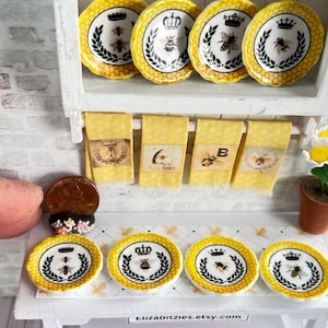 May include: Set of six miniature yellow and black bee themed plates with a crown design. The plates are displayed on a white shelf with yellow and black patterned towels. The plates are approximately the size of a US quarter.