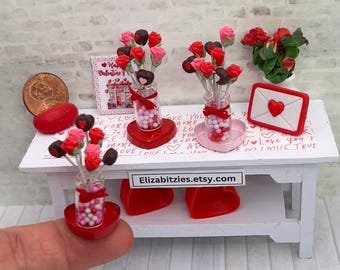 May include: Two miniature Valentine's Day bouquets with red and pink roses and chocolate hearts on sticks in glass jars filled with candy hearts. The bouquets are on a white table with a red heart-shaped base and a pink heart-shaped base. The table has a sign that says "Elizabitzies.etsy.com".