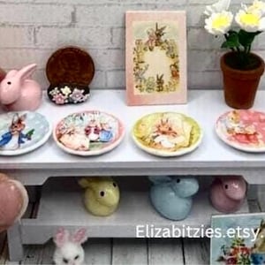 May include: A white miniature table with two shelves. The top shelf has four small plates with bunny illustrations, a pink bunny figurine, and a brown decorative piece. The bottom shelf has three bunny figurines in yellow, blue, and pink. A small white flower pot with white flowers is on the table.